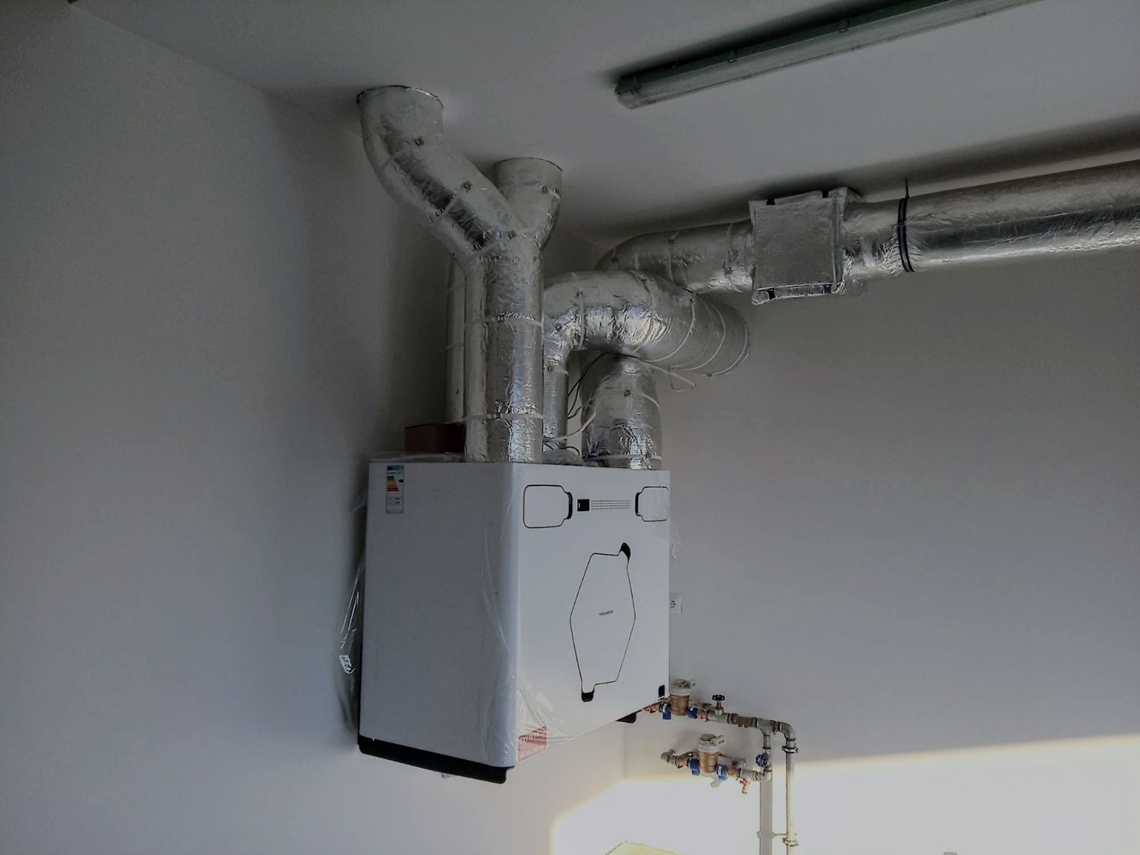 Biała pompa ciepła zamontowana na ścianie, z widocznymi, zaizolowanymi rurami wentylacyjnymi. Kompaktowa instalacja w jasnym pomieszczeniu.