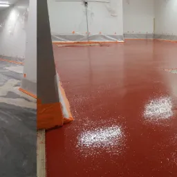 Etap prac: Por&oacute;wnanie surowej i pomalowanej na czerwono posadzki żywicznej w pomieszczeniu przemysłowym, z pomarańczową taśmą zabezpieczającą ściany.