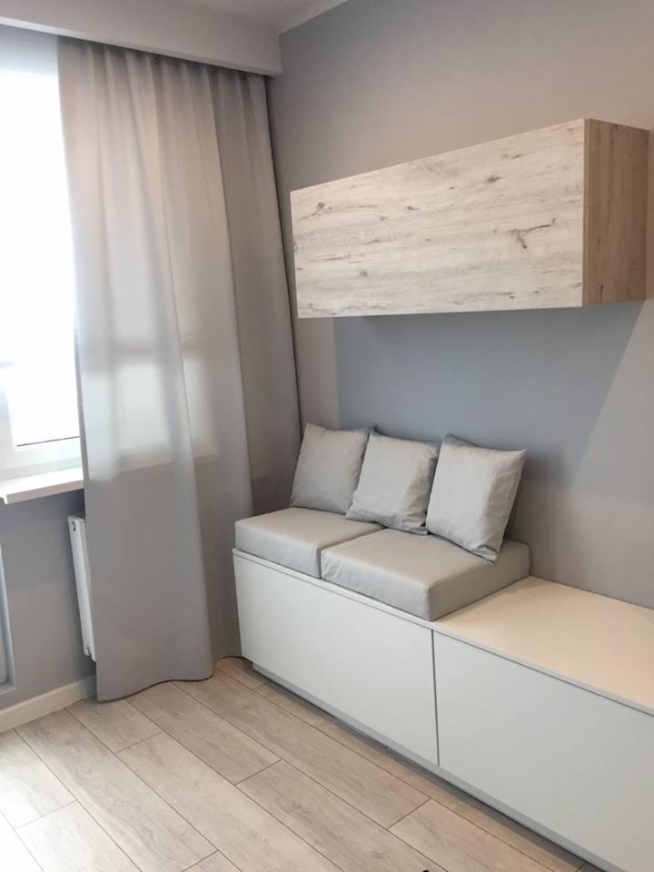 Jasnoszara, minimalistyczna aranżacja wnętrza z siedziskiem z szarymi poduszkami, białym meblem z szafkami i drewnopodobnym regałem wiszącym, uzupełniona przez zwiewne, jasne zasłony wpadające...