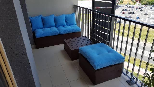 Balkon z drewnianymi meblami ogrodowymi z błękitnymi poduszkami, widok na parking i zieleń za barierką.