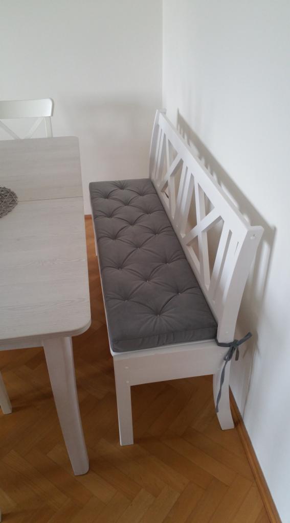 Biała ławka z drewnianym oparciem w kształcie krzyża, z szarym, pikowanym siedziskiem, dopasowana do stołu w jasnym odcieniu drewna.