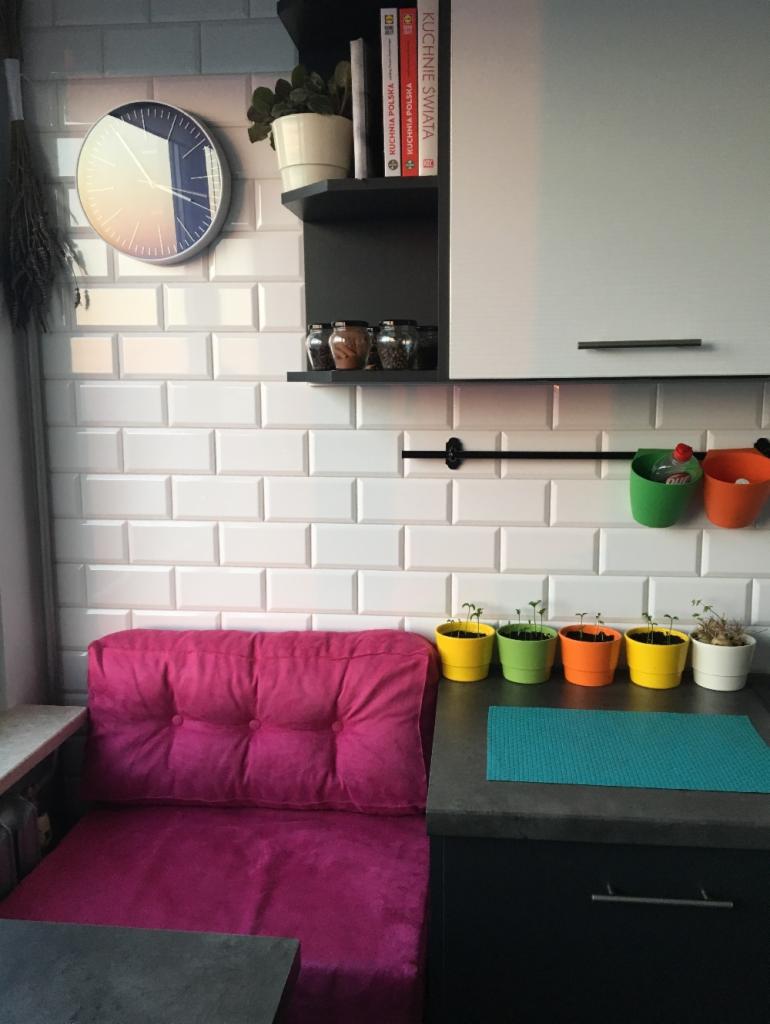 Kącik kuchenny z fuksjową, pikowaną poduszką na siedzisku, białymi płytkami metra na ścianie, zegarem z motywem zachodu słońca i rzędem kolorowych doniczek z sadzonkami.