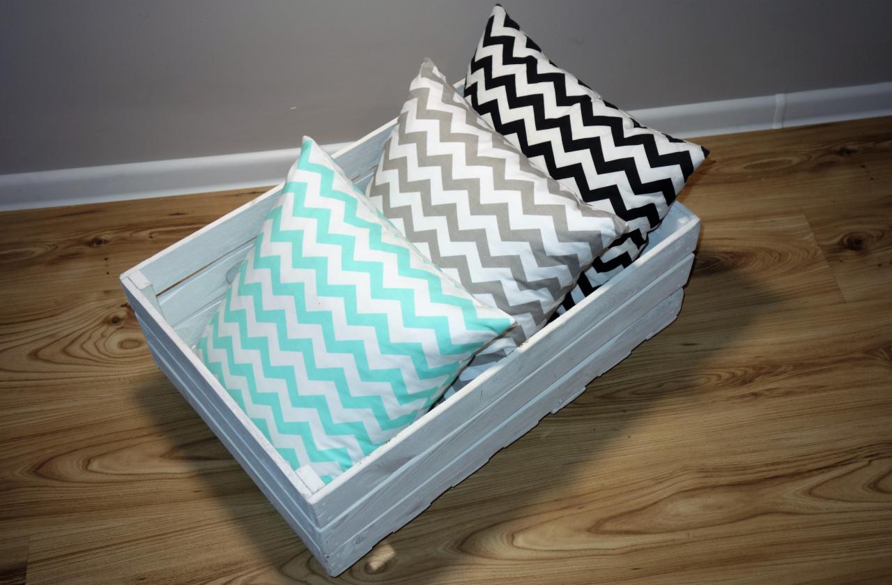 Trzy poduszki dekoracyjne w drewnianej, pomalowanej na biało skrzynce: turkusowa, szara i czarna, wszystkie w zygzakowaty wzór, ułożone na drewnianej podłodze.