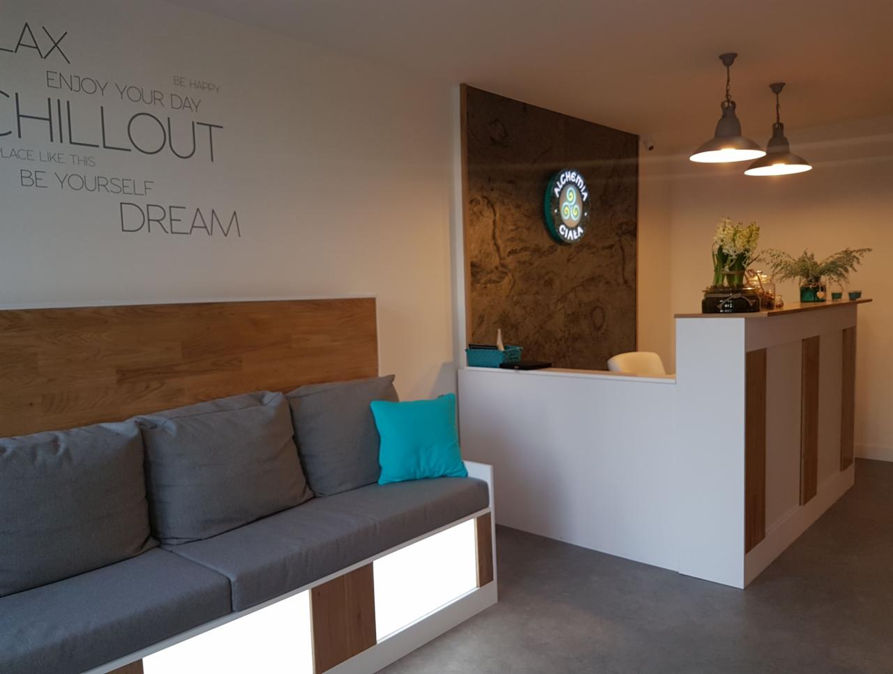 Wnętrze salonu z recepcją: szara sofa z drewnianym zagłówkiem i turkusową poduszką, biała lada recepcyjna z drewnianymi wstawkami, na ścianie logo firmy i napisy motywacyjne, oświetlenie lampami...