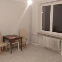 Jasne wnętrze pokoju z dwoma krzesłami i stołem, widoczne panele podłogowe i grzejnik pod oknem.