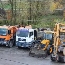 Rząd pojazdów budowlanych: biały Renault, pomarańczowy Mercedes-Benz, biały MAN, żółta ładowarka i koparka Liebherr, stojących na placu budowy z ziemią w tle.