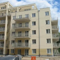 Nowoczesny budynek wielorodzinny z balkonami, świeżo po tynkowaniu i malowaniu elewacji w odcieniach beżu i żółci, widoczne materiały budowlane i przygotowane podłoże.