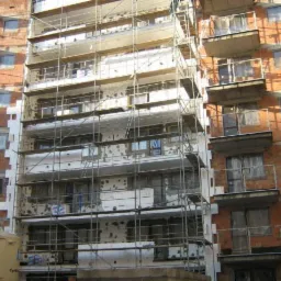 Budynek wielorodzinny w trakcie prac: częściowo ocieplona elewacja, widoczne rusztowania i zbiorniki z zaprawą, surowa cegła na sąsiedniej części budynku.