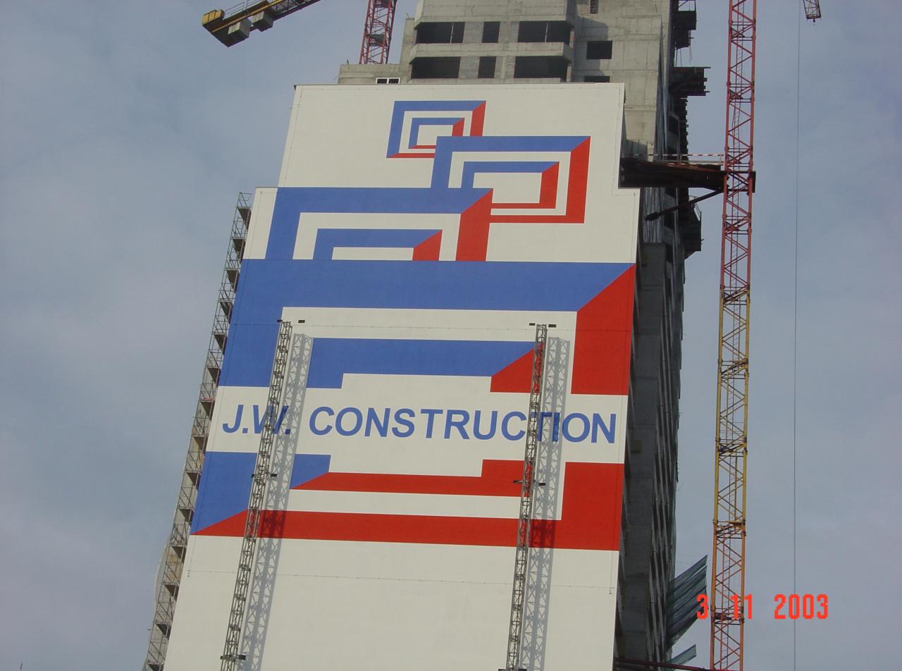 Wysoki budynek w trakcie budowy z pomalowaną elewacją w geometryczne wzory w kolorach czerwonym i niebieskim oraz napisem 'J.W. CONSTRUCTION'. Widoczne rusztowania i żurawie budowlane.