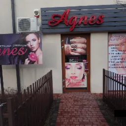Fasada salonu kosmetycznego Agnes z szyldem, plakatem z wizerunkiem kobiety z kwiatem i informacjami o ofercie, drzwi wejściowe z naklejonymi zdjęciami paznokci i makijażu, oraz listą zabiegów.