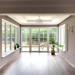 ROS-BUD Łukasz Rosiński - Jasne wnętrze z dużymi oknami z widokiem na zieleń, drewniana podłoga, ściany w odcieniach szarości z dekoracyjnymi listwami, sufit z oświetleniem i rośliny doniczkowe na parapetach.