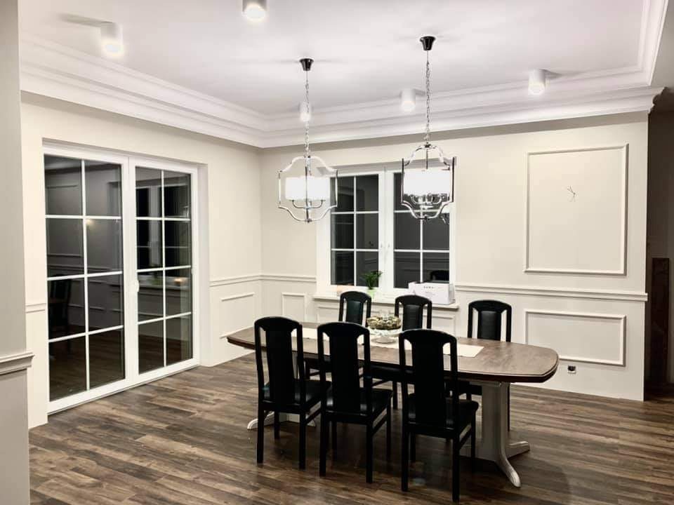 Elegancka jadalnia w odcieniach beżu z drewnianą podłogą, białymi panelami ściennymi i listwami przysufitowymi, oświetlona wiszącymi lampami i punktowymi reflektorami, z dużym oknem i przeszklonymi...