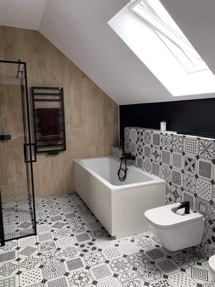 Nowoczesna łazienka na poddaszu z białą wanną, wiszącą toaletą, czarną armaturą i wzorzystymi płytkami podłogowymi oraz ściennymi, doświetlona oknem dachowym.