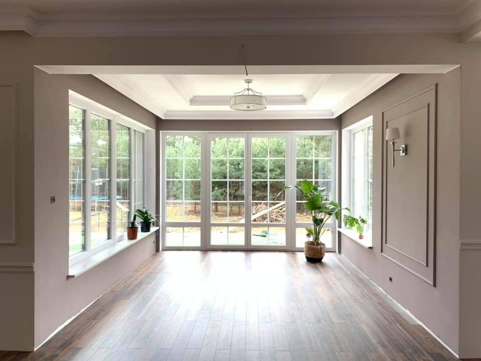 Jasne wnętrze z dużymi oknami z widokiem na zieleń, drewniana podłoga, ściany w odcieniach szarości z dekoracyjnymi listwami, sufit z oświetleniem i rośliny doniczkowe na parapetach.