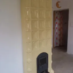 Żółty, kaflowy piec z czarnymi drzwiczkami, widoczny w rogu pokoju z surowym wykończeniem i drewnianą belką stropową, dekoracja w kształcie księżyca na ścianie.