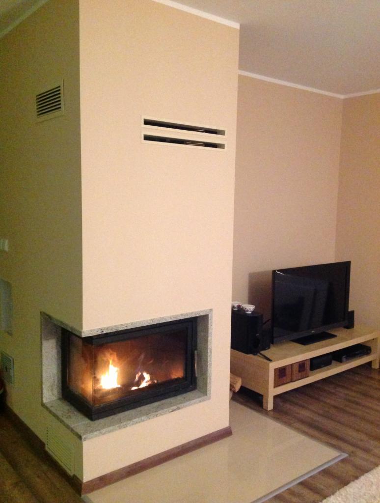 Narożny kominek z czarną ramą i płomieniami w jasnym salonie z beżowymi ścianami i drewnianą podłogą. Nad kominkiem widoczne dwa poziome otwory wentylacyjne.