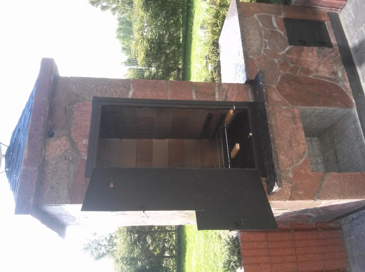 Otwarty piec ogrodowy z kamienia z metalowymi drzwiczkami i rusztem wewnątrz, na tle zieleni.