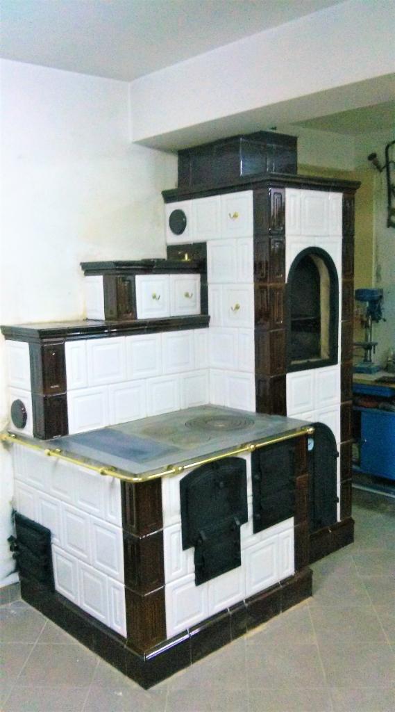 Wielopoziomowy piec kaflowy z białymi i brązowymi kaflami, z metalowymi drzwiczkami i płytą grzewczą, widoczny w pomieszczeniu z jasnymi ścianami.