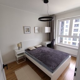 Twoi Wykonawcy Tomasz Przewłocki - Nowoczesna sypialnia w jasnych barwach z drewnianą podłogą, białym łóżkiem, stolikiem nocnym i okrągłą lampą sufitową. Za oknem widoczny wysoki budynek mieszkalny.