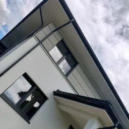 Nowoczesna elewacja domu w Białymstoku z widocznymi oknami z czarnymi ramami i systemem odprowadzania wody, sfotografowana pod kątem w pochmurny dzień.