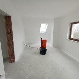 Twoi Wykonawcy Tomasz Przewłocki - Białe, świeżo pomalowane wnętrze pokoju z oknem dachowym i standardowym oknem, widoczny grzejnik elektryczny i wiadro, ślady farby na podłodze, przygotowania do dalszych prac wykończeniowych.