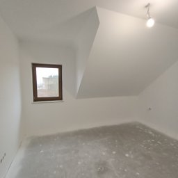 Twoi Wykonawcy Tomasz Przewłocki - Poddasze po szpachlowaniu, z widocznym oknem w drewnianej ramie i surową podłogą przygotowaną do dalszych prac wykończeniowych.