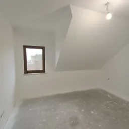 Poddasze po szpachlowaniu, z widocznym oknem w drewnianej ramie i surową podłogą przygotowaną do dalszych prac wykończeniowych.