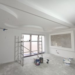 Twoi Wykonawcy Tomasz Przewłocki - Wnętrze w trakcie prac wykończeniowych: pomalowane ściany na biało, dekoracyjny sufit z gładzią, aluminiowy ruszt, wiaderka z farbą oraz wiertarka akumulatorowa na zabezpieczonej podłodze.