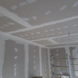 Wnętrze w trakcie prac wykończeniowych: ściany i sufit pokryte płytami kartonowo-gipsowymi, widoczne łączenia szpachlowane, drabina aluminiowa, rolka papieru ściernego przy ścianie.