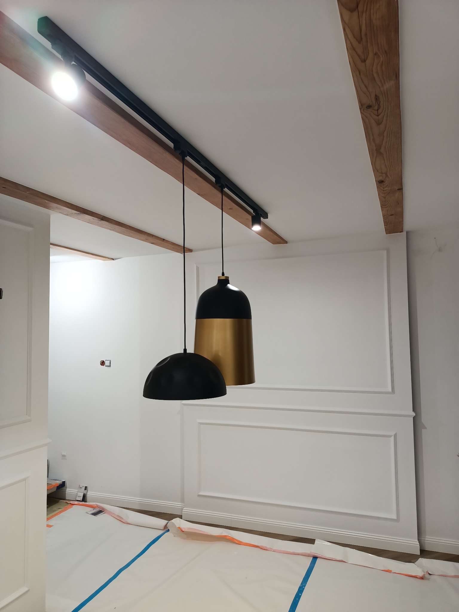 Wnętrze pokoju w trakcie remontu: ściany z dekoracyjnymi listwami, drewniane belki na suficie z zamontowanym oświetleniem szynowym i wiszącymi lampami w kolorach czerni i złota, podłoga...