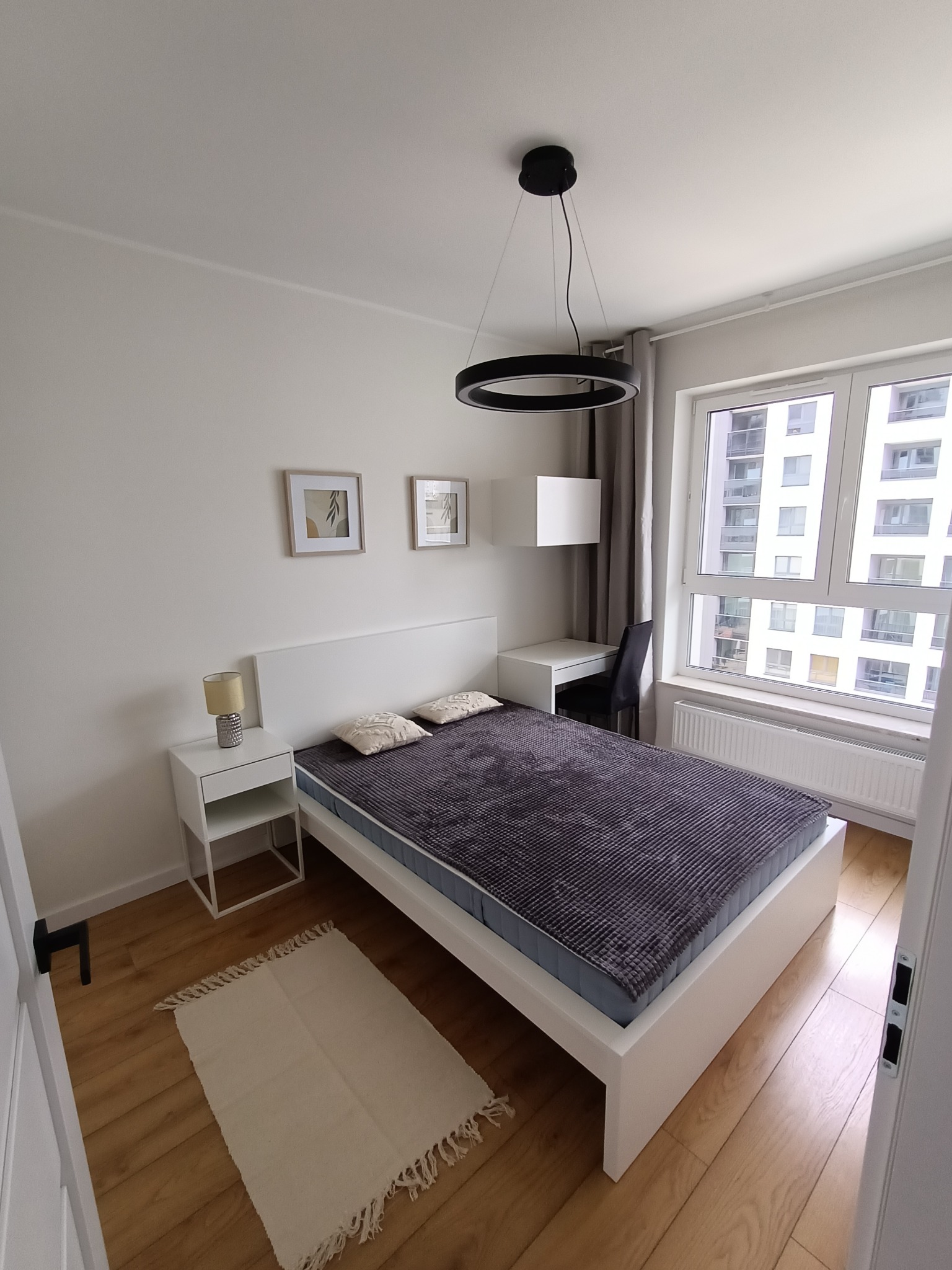 Nowoczesna sypialnia w jasnych barwach z drewnianą podłogą, białym łóżkiem, stolikiem nocnym i okrągłą lampą sufitową. Za oknem widoczny wysoki budynek mieszkalny.