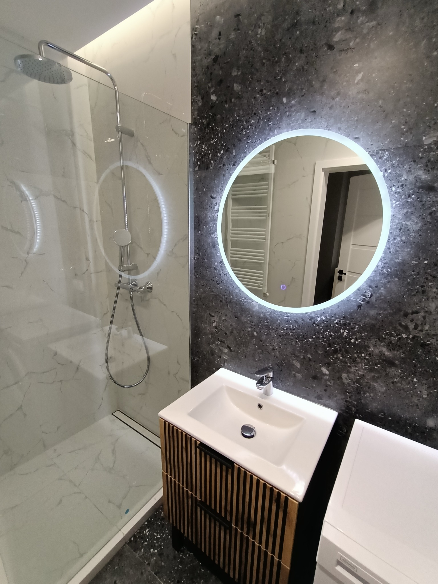 Nowoczesna łazienka z kabiną prysznicową typu walk-in, umywalką nablatową z szafką z drewnianymi lamelami i okrągłym lustrem z podświetleniem LED na tle ściany z ciemnego lastryko.