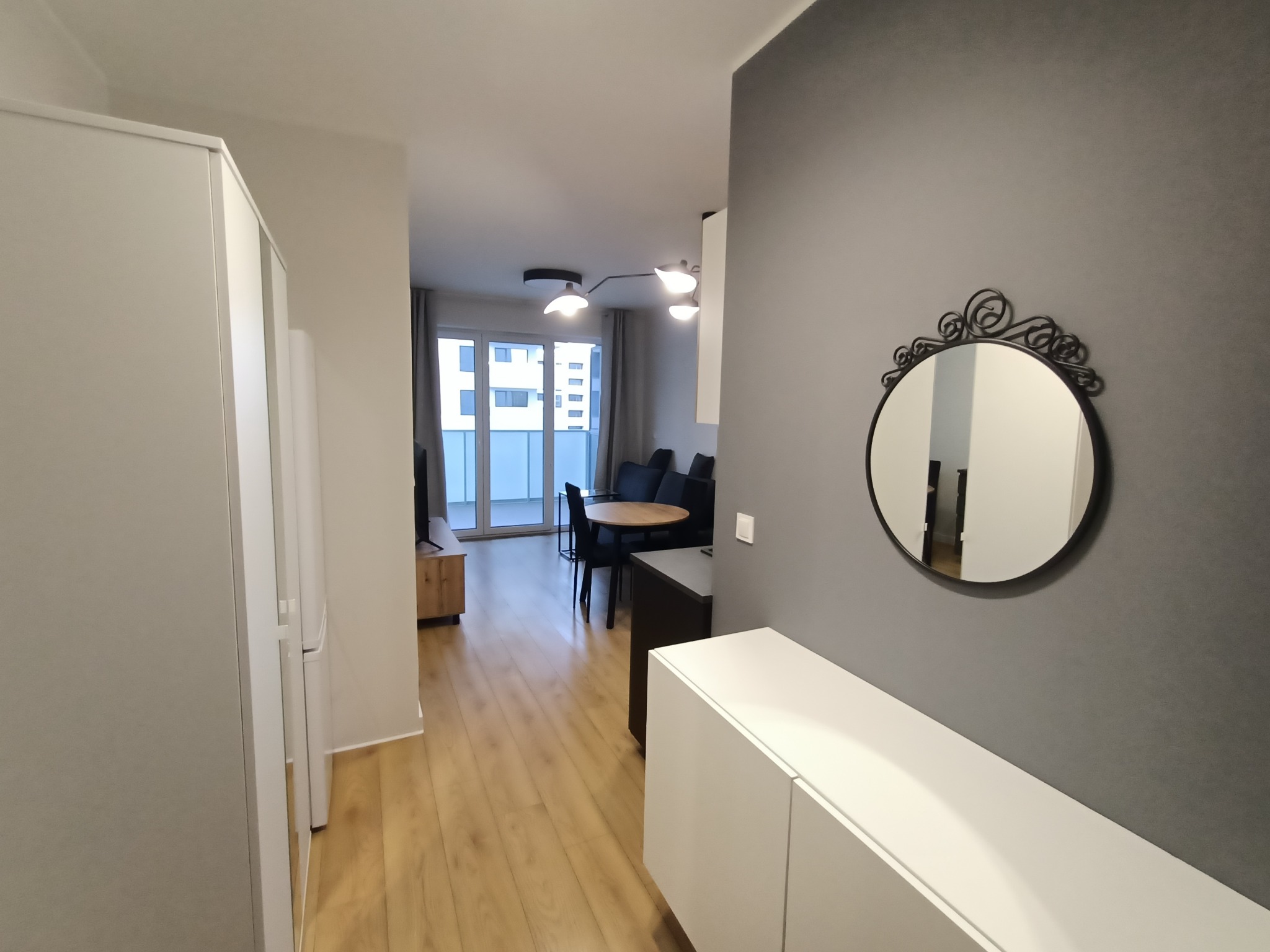 Nowoczesne wnętrze mieszkania z szarą ścianą, okrągłym lustrem z ozdobną ramą, białą komodą, drewnianą podłogą i widokiem na salon z okrągłym stołem i balkonem.