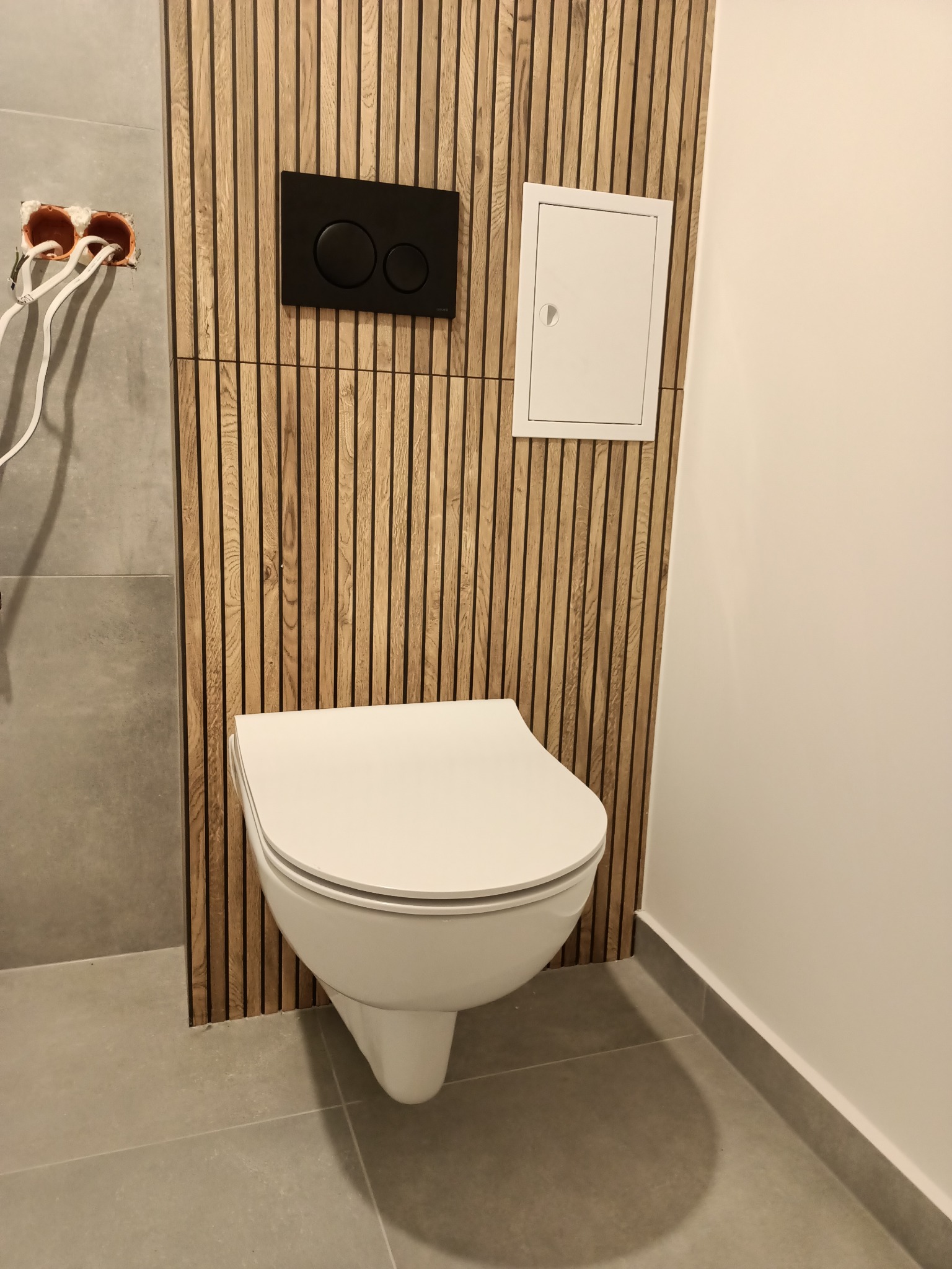 Podwieszana miska WC na tle drewnianej ściany z pionowych listewek, z czarnym przyciskiem spłukującym i białym włazem rewizyjnym; widoczne niedokończone przyłącze elektryczne.