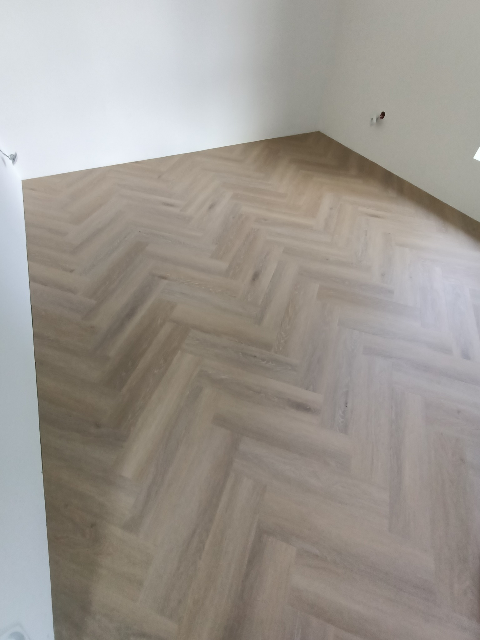 Ułożona podłoga z paneli w jodełkę francuską w jasnym odcieniu drewna, widok z góry na fragment pokoju z białymi ścianami i widocznym gniazdkiem elektrycznym.