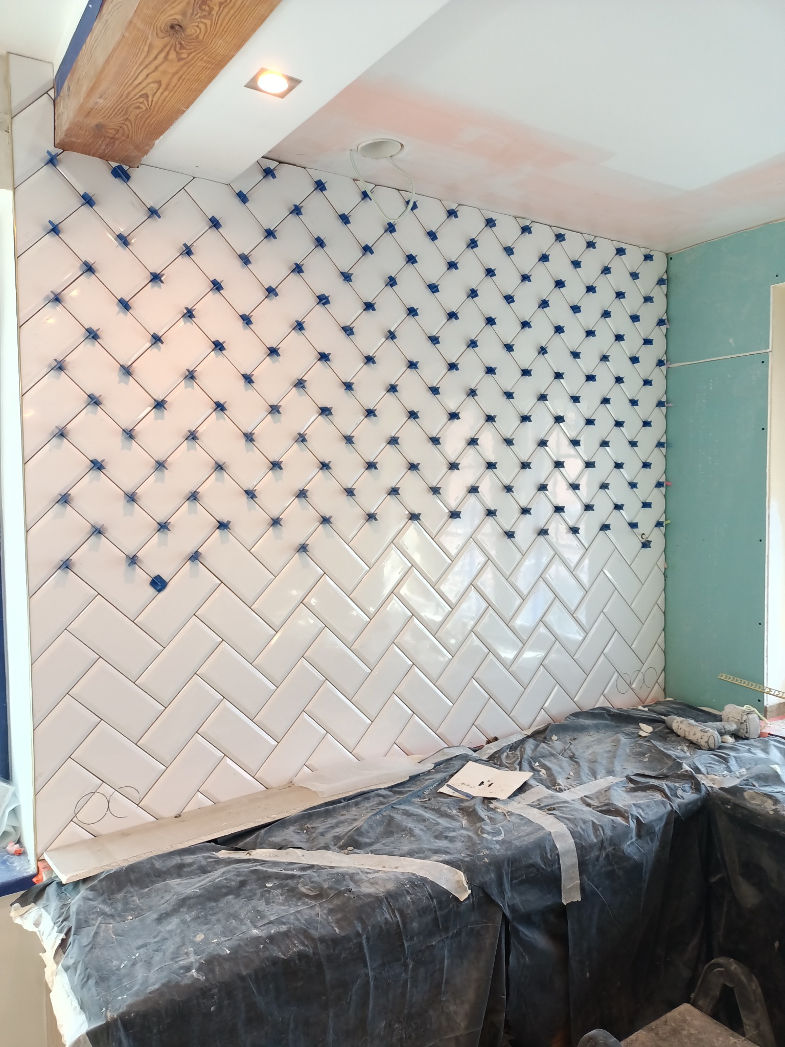 Białe, błyszczące płytki ułożone w jodełkę na ścianie, z niebieskimi krzyżykami dystansowymi, częściowo przykryte folią ochronną, widoczna płyta gipsowo-kartonowa na sąsiedniej ścianie.