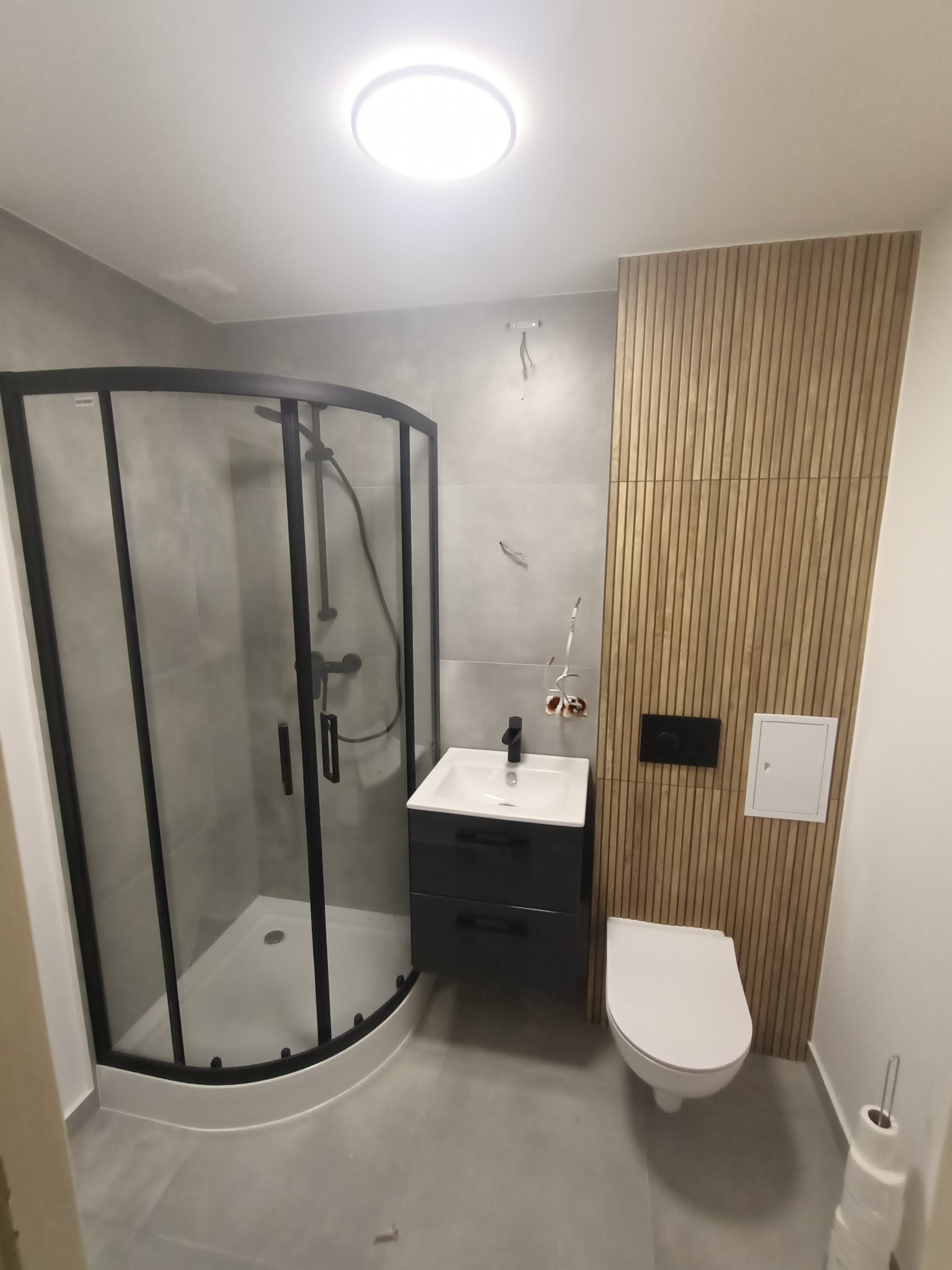 Nowoczesna łazienka z kabiną prysznicową z czarnymi ramami, szafką pod umywalką w kolorze grafitowym, drewnianą okładziną ścienną z pionowych paneli oraz podwieszaną toaletą.