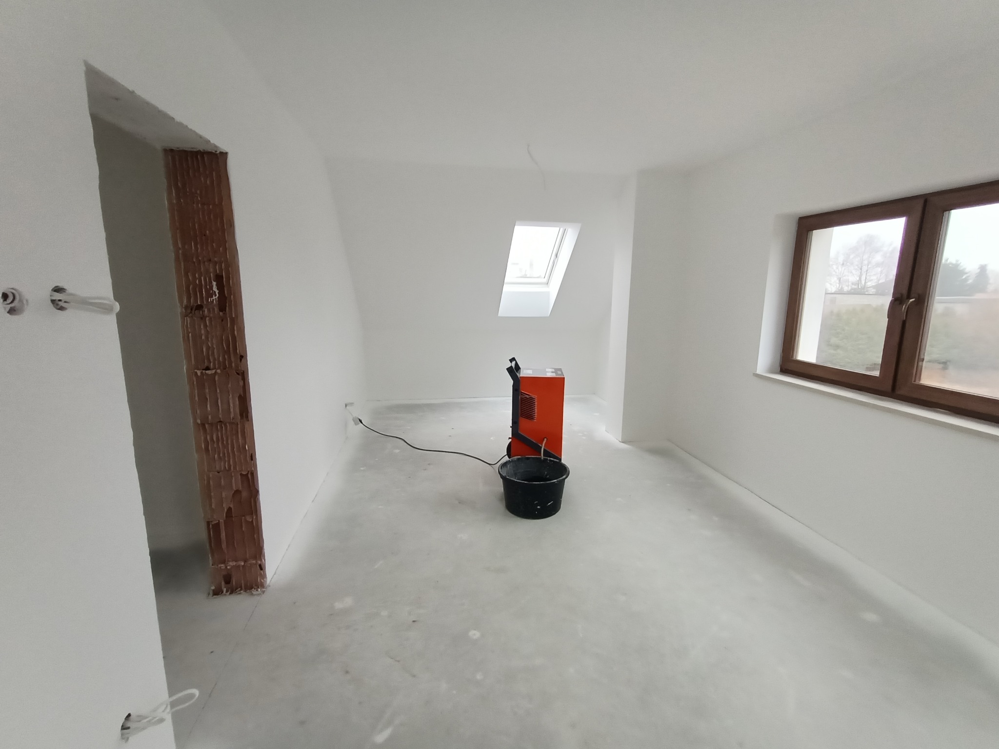 Białe, świeżo pomalowane wnętrze pokoju z oknem dachowym i standardowym oknem, widoczny grzejnik elektryczny i wiadro, ślady farby na podłodze, przygotowania do dalszych prac wykończeniowych.