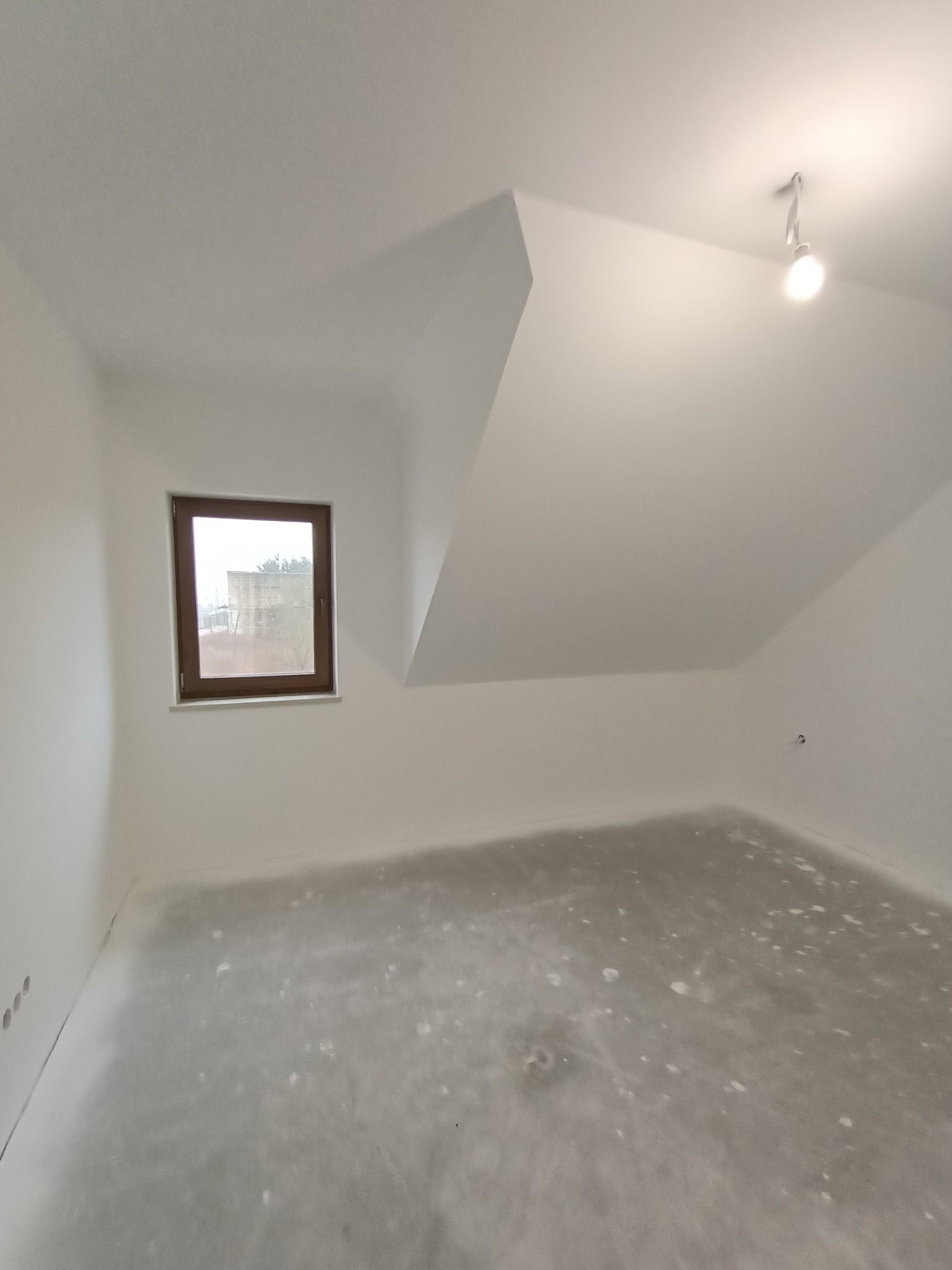 Poddasze po szpachlowaniu, z widocznym oknem w drewnianej ramie i surową podłogą przygotowaną do dalszych prac wykończeniowych.