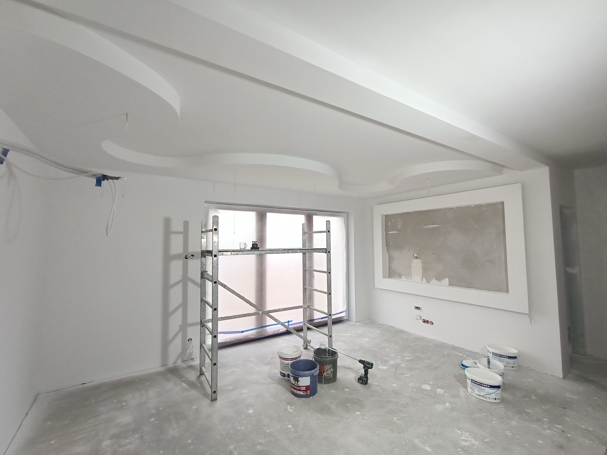 Wnętrze w trakcie prac wykończeniowych: pomalowane ściany na biało, dekoracyjny sufit z gładzią, aluminiowy ruszt, wiaderka z farbą oraz wiertarka akumulatorowa na zabezpieczonej podłodze.