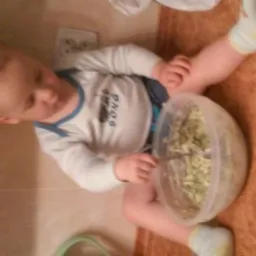 Dziecko siedzi na podłodze, trzymając miskę z sałatką i łyżką. Ubrany w białą koszulkę z niebieskim wykończeniem. Widok z góry, lekko obrócony.