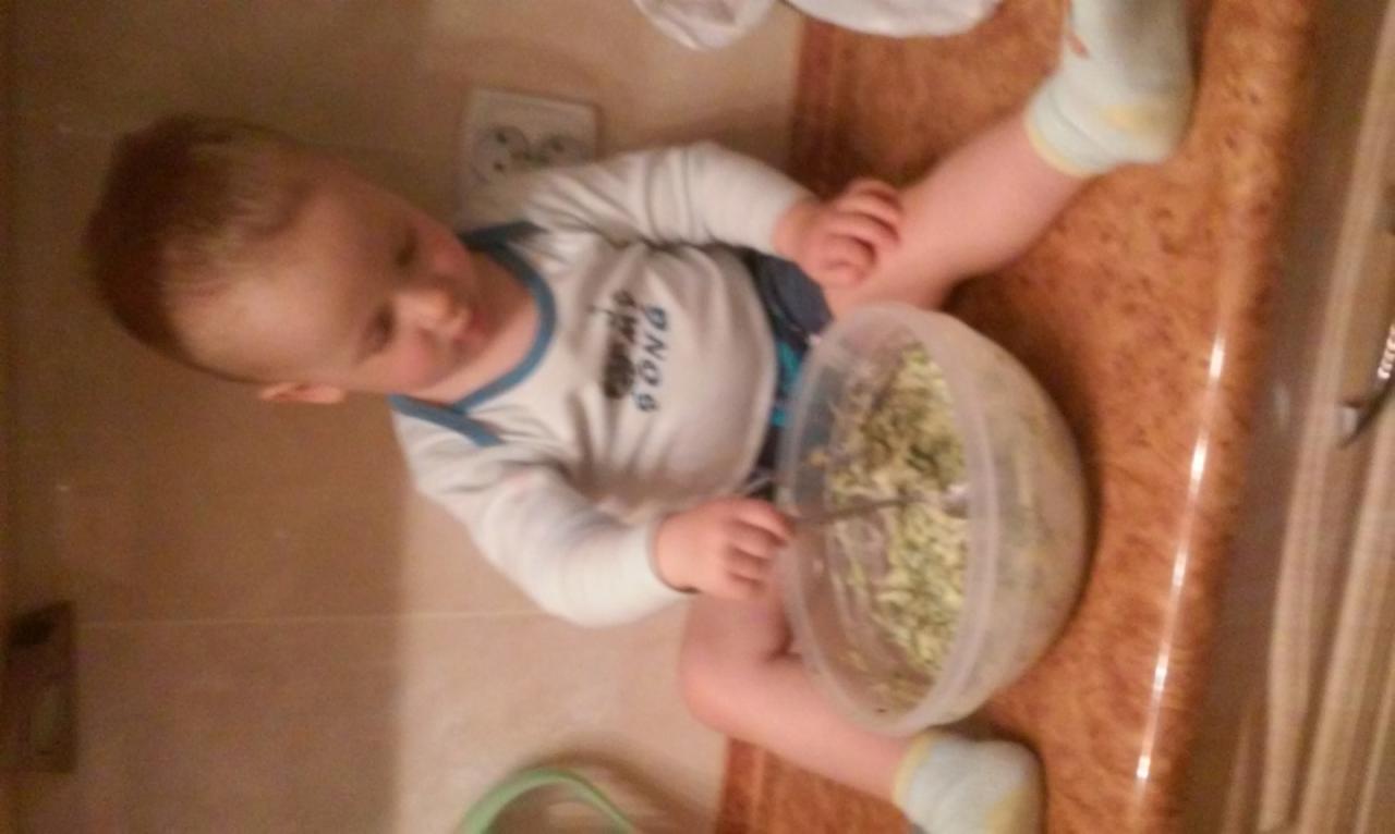 Dziecko siedzi na podłodze, trzymając miskę z sałatką i łyżką. Ubrany w białą koszulkę z niebieskim wykończeniem. Widok z góry, lekko obrócony.