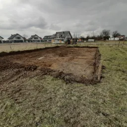 Przygotowanie terenu pod budowę: wykopany fundament pod budynek jednorodzinny z widocznym traktorem w tle i okoliczną zabudową.
