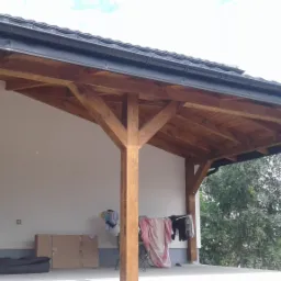 Drewniana konstrukcja zadaszenia tarasu z czarnymi rynnami, widok z dołu na drewniane belki i elementy wsporcze, w tle ściana budynku z dekoracyjną okładziną imitującą kamień, na tarasie suszące...