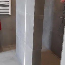 Nowoczesna łazienka z szarymi płytkami, podświetlanym pionowym panelem i grzejnikiem drabinkowym, widok na kabinę prysznicową z deszczownicą.