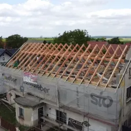 Dom jednorodzinny w trakcie wymiany konstrukcji dachu, widoczne drewniane krokwie i łaty, rusztowania z siatką ochronną z reklamą Weber i Sto, w tle inne domy i pola uprawne.