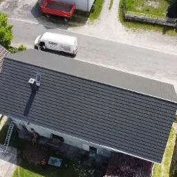 Widok z góry na dom z ciemnoszarą dachówką, białą obudową komina i nowo wykończonym dachem; w tle biały van firmy budowlanej i czerwona przyczepa.