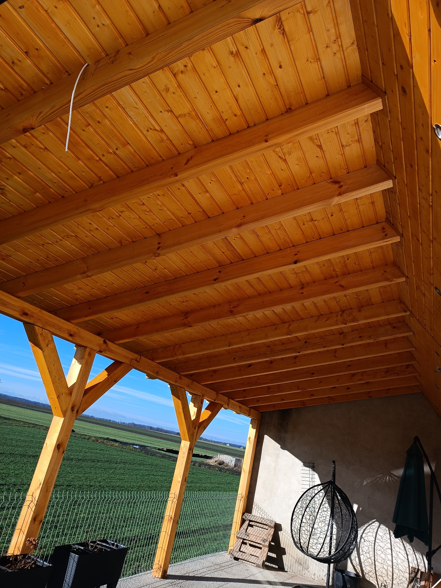 Drewniana konstrukcja zadaszenia tarasu z widokiem na zielone pole i błękitne niebo, w tle ściana budynku z wiszącym fotelem i parasolem.