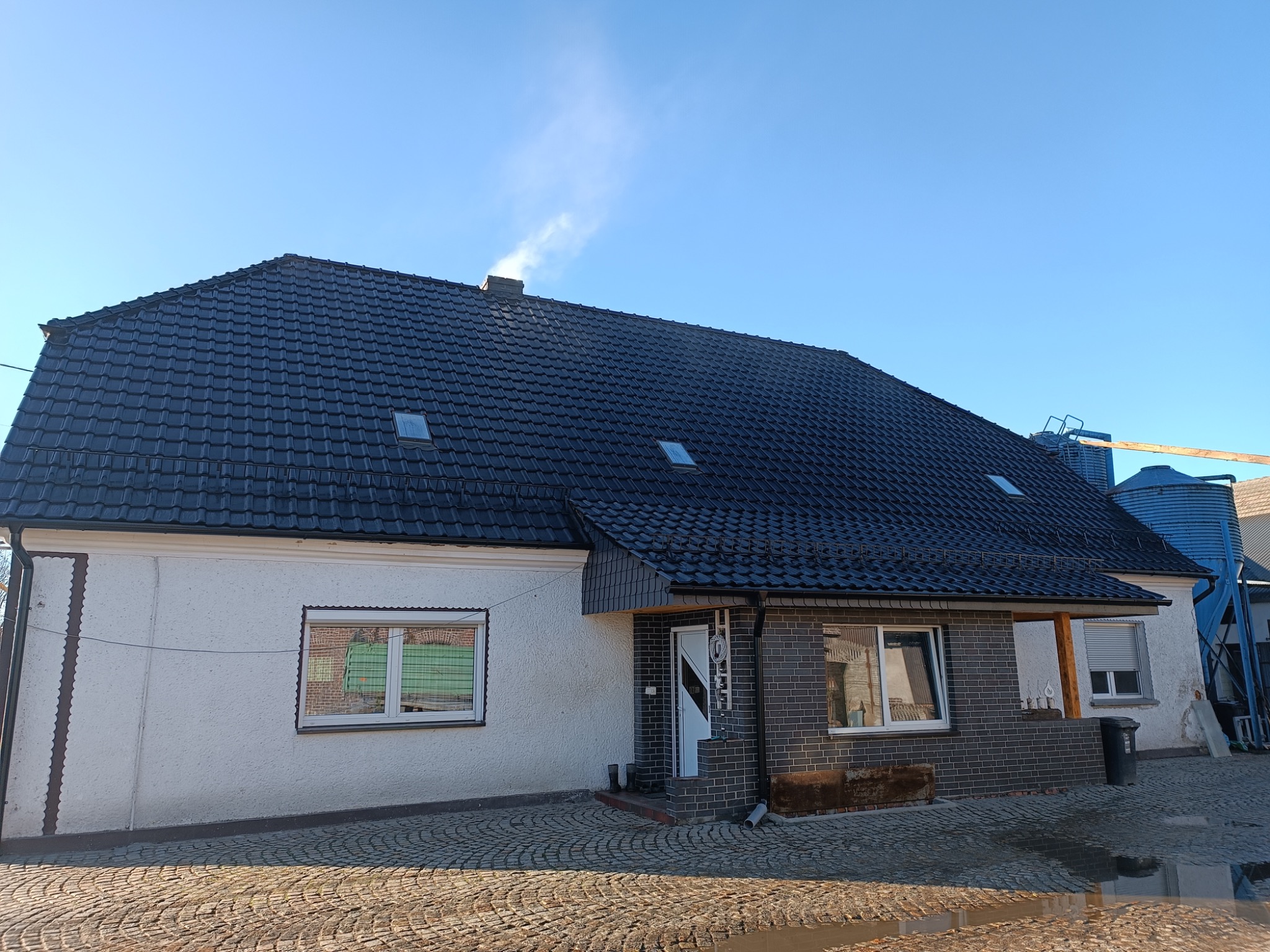 Dom jednorodzinny z nowym, czarnym dachem z dachówki ceramicznej, widoczne okna dachowe i komin z wydobywającym się dymem, elewacja częściowo w kolorze białym, częściowo w odcieniach szarości, bruk...