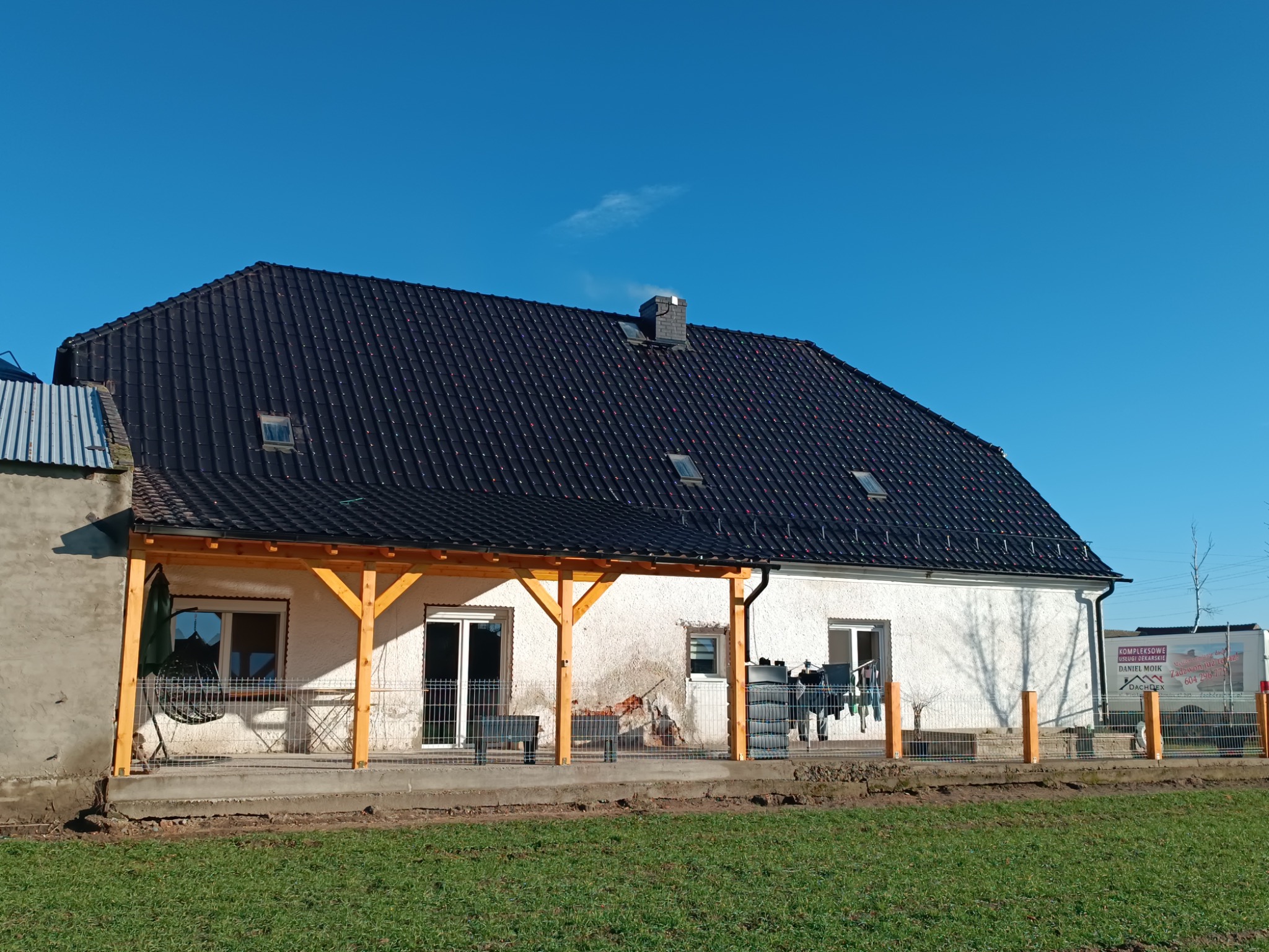 Dom z nowym, czarnym dachem z dachówki ceramicznej, widoczne okna dachowe i komin, jasna elewacja, drewniana konstrukcja zadaszenia tarasu w trakcie budowy, ogrodzenie z metalowej siatki, błękitne...